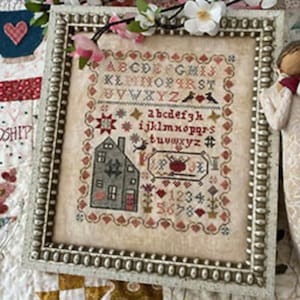 May include: Framed cross-stitch sampler with a house, alphabet, numbers, and heart motifs. The sampler is in a silver-colored frame and is decorated with pink and white flowers. The background is a patchwork quilt.