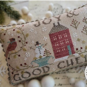 May include: A cross-stitch sampler with a beige background featuring a red house, a snowman, and the text "Joy & Good Cheer".