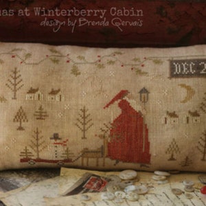 May include: A beige pillow with a cross-stitch design depicting a winter scene with a red-robed figure, a snowman, and houses. The text "DEC 25" is stitched on the pillow. The pillow is on a wooden surface with buttons and a piece of paper.