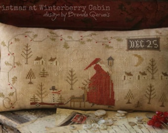 May include: A beige pillow with a cross-stitch design depicting a winter scene with a red-robed figure, a snowman, and houses. The text "DEC 25" is stitched on the pillow. The pillow is on a wooden surface with buttons and a piece of paper.