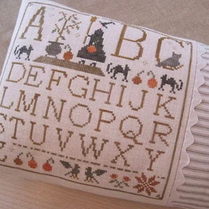 May include: A decorative pillow with a cross-stitch alphabet design. The design includes Halloween-themed motifs such as a witch, black cats, pumpkins, and bats. The pillow has a striped fabric border with a scalloped edge.