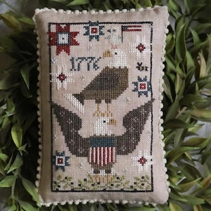 May include: A rectangular, beige, cross-stitched pillow featuring a patriotic design. The design includes an eagle with outstretched wings, the number "1776", stars, and a small American flag. The pillow has a cream-colored pom-pom trim.