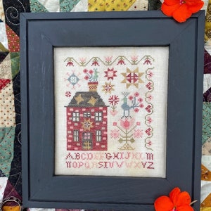 May include: A framed cross-stitch embroidery featuring a red house with a chimney, a bird, and a flower garden. The alphabet is stitched below the house. The frame is black and the background is white.