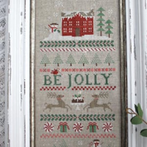 May include: Framed cross-stitch artwork with a winter holiday theme. The design features a red house, trees, reindeer, and the words "BE JOLLY." The artwork is framed in a distressed white wooden frame.