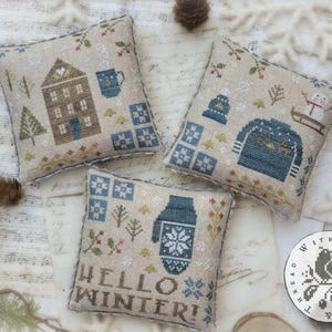 May include: Three square decorative pillows with cross-stitch winter designs. One pillow features a house and mug, another a sweater and hat, and the third says "Hello Winter" with a mitten. The pillows are beige with blue and green accents.