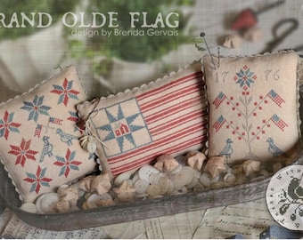 May include: Three decorative patriotic pillows with embroidered designs. One pillow features a star and stripes pattern, another displays a star and house design, and the third has a tree with flags and the number "1776". The pillows are in a metal tray with buttons.