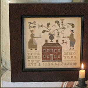 May include: A framed cross-stitch sampler with a house, a plant, two figures, and a dog. The sampler includes the alphabet, numbers, and the words "The Candlestick Maker Sampler" and "design by Brenda Gervais".