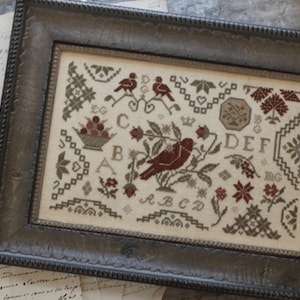 May include: Framed cross-stitch sampler with a dark wooden frame. The sampler features a central bird motif, floral and geometric patterns, and the text "QUAKER HANDEWORK design by Brenda Gervais". The color palette includes shades of red, brown, and beige.