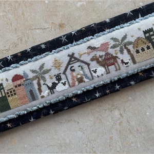 May include: A cross-stitch sampler depicting the nativity scene with a star, a camel, palm trees, and a small village. The scene is stitched on a white linen fabric and framed with a blue and white trim.