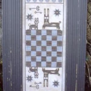 May include: Framed artwork featuring a cross-stitch design with a checkerboard pattern in blue and brown. The artwork includes stylized rabbits, keys, and star motifs. The frame is a distressed dark gray with a ribbed texture.