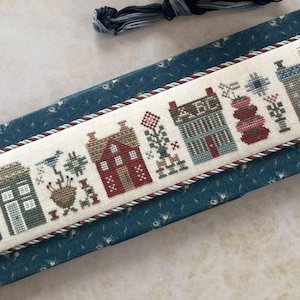 May include: A cross-stitch sampler featuring a row of stylized houses in various colors, including blue, red, and green. The houses are decorated with floral and geometric patterns. The sampler is framed with a twisted cord and rests on a blue patterned surface.