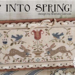 May include: Cross-stitch design with the text "HOP INTO SPRING!" featuring two brown rabbits, blue birds, and a red tulip. The design is framed in a light-colored, scalloped edge with a cream-colored bow.