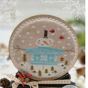 May include: A cross-stitch pattern of a snowman wearing a blue sweater with snowflakes, standing in a snowy forest with red birds flying overhead. The design is framed in a round wooden hoop and the text "SNOW MAGICAL! design by Brenda Gervais" is at the top of the image.