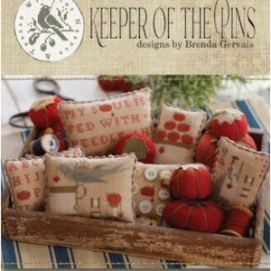 May include: A collection of 10 small, handmade pincushions in a wooden box. The pincushions are made of fabric and have various designs, including a bird, a heart, and a needle. The pincushions are filled with stuffing and have a red, white, and blue color scheme. The box is made of wood and has a rustic look. The text "KEEPER OF THE PINS" is printed on the box.