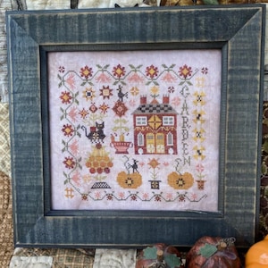 May include: A framed cross-stitch embroidery featuring a red house with a black cat and pumpkins. The embroidery is surrounded by a floral pattern and the text "Garden" is stitched vertically on the right side of the house.