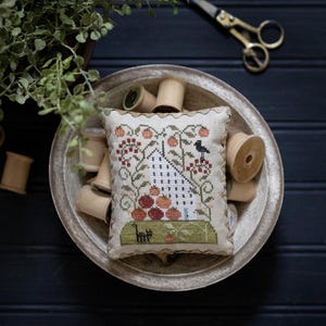 May include: A white and green cross-stitch pillow with a house, trees, and a bird. The pillow is surrounded by spools of thread in a bowl.