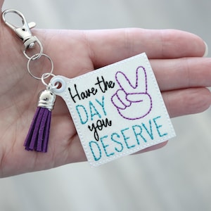 May include: White square keyring with a purple tassel. The keyring has the text "Have the day you deserve" and a purple peace sign embroidered on it.