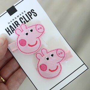 May include: Two pink pig hair clips with black eyes and a pink nose. The clips are on a white card with the text "Handmade Hair Clips".