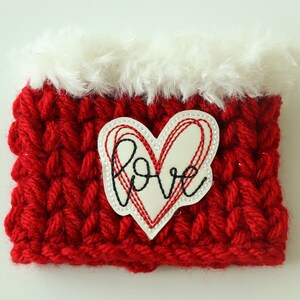 May include: Red crocheted cup cozy with a white faux fur trim and a white heart-shaped patch with the word "love" embroidered in black and red thread.