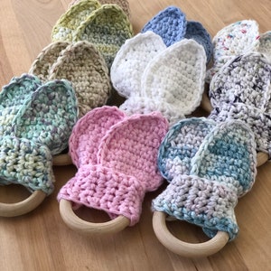 May include: A set of ten crocheted baby teethers in various colors, including green, blue, pink, white, and beige. Each teether has a wooden ring attached.