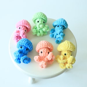 May include: A set of six crocheted octopus plushies in various pastel colors, including pink, blue, green, yellow, and orange. The plushies are arranged on a white pedestal.