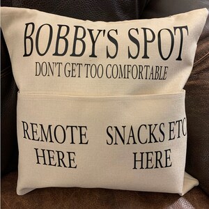 May include: Beige pillow with black text that reads "BOBBY'S SPOT DON'T GET TOO COMFORTABLE" above a pocket with the text "REMOTE HERE" and "SNACKS ETC HERE". The pillow is on a brown surface.