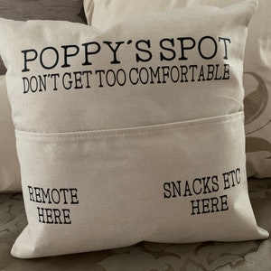 May include: Beige pillow with a pocket, featuring the text "POPPY'S SPOT DON'T GET TOO COMFORTABLE". Below the pocket, it says "REMOTE HERE" on the left and "SNACKS ETC HERE" on the right. The pillow is on a patterned surface.