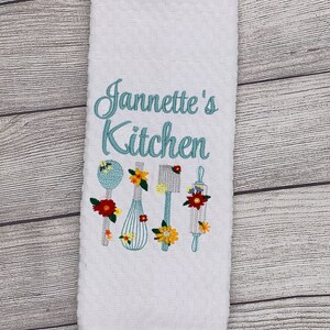 May include: White kitchen towel with blue embroidery that says "Jannette's Kitchen" and features a whisk, rolling pin, spoon, and spatula with floral accents.