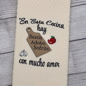 May include: A white kitchen towel with the text "En Esta Cocina hay Sazon Adobo Sofrito con mucho amor" embroidered in black. The embroidery also includes a brown cutting board, two cloves of garlic, and a red tomato.