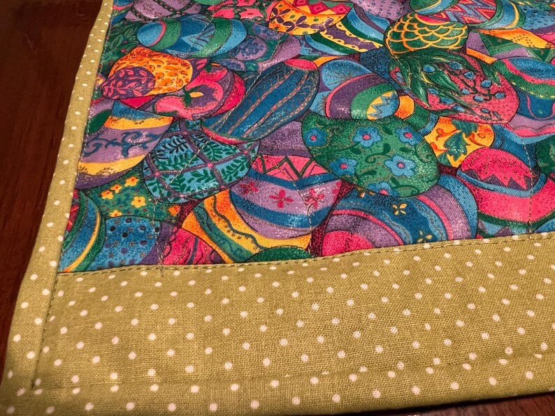 Puede incluir: Un camino de mesa verde con lunares blancos. El camino de mesa tiene un borde colorido con un patr&oacute;n de huevos de Pascua.