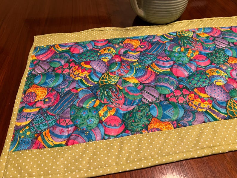 Puede incluir: Un camino de mesa verde con lunares blancos presenta un patr&oacute;n colorido de huevos de Pascua. Los huevos est&aacute;n decorados con varios dise&ntilde;os y colores, incluyendo morado, azul, verde, amarillo y rosa.