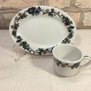 May include: White ceramic oval serving platter and matching teacup. Both pieces feature a decorative border of green leaves, blue and brown flowers. The teacup has a small handle. The set is displayed on a white surface.