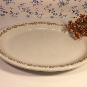 May include: Off-white oval serving platter with a decorative gold border. The border features a repeating geometric pattern. The platter is empty and sits on a white surface. Dried orange flowers are in the background.