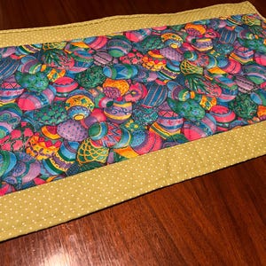 Puede incluir: Un camino de mesa verde con un borde de lunares blancos. El centro del camino de mesa presenta un patr&oacute;n colorido de huevos de Pascua.