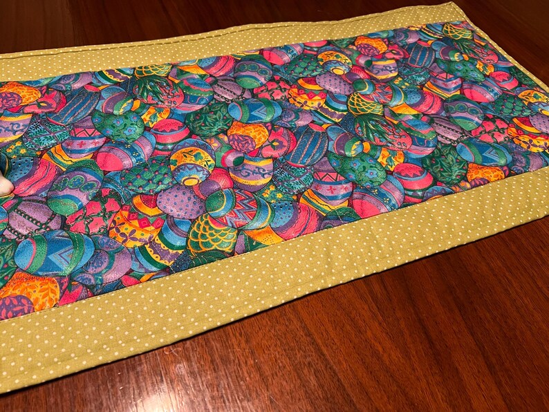 Puede incluir: Un camino de mesa verde con un borde de lunares blancos. El centro del camino de mesa presenta un patr&oacute;n colorido de huevos de Pascua.