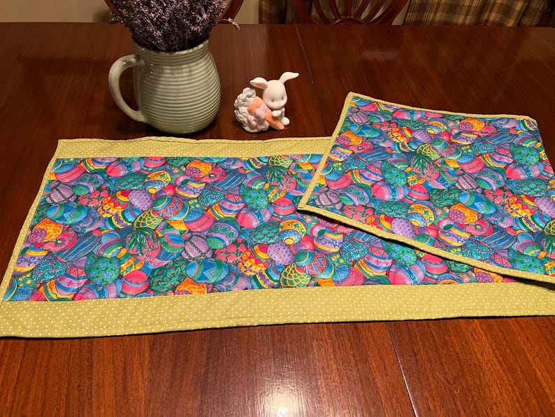 Puede incluir: Dos caminos de mesa coloridos con un patr&oacute;n de huevos de Pascua. Los caminos de mesa son verdes, azules, morados y amarillos. Los caminos de mesa tienen un borde amarillo con lunares blancos.