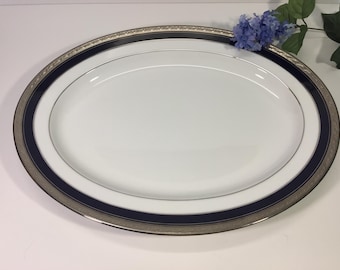 Op de afbeelding: Een ovale serveerschaal van porselein met een helder wit midden, een brede marineblauwe rand en een buitenrand versierd met een gedetailleerd zilverkleurig metalen patroon. De schaal heeft een gladde, glanzende afwerking en een symmetrische vorm.