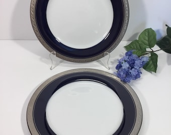 Op de afbeelding: Twee ronde dinerborden met een wit midden, een brede marineblauwe band en een zilverkleurige rand met een ingewikkeld reliëfpatroon. De borden zijn cirkelvormig met een gladde afwerking en een opvallend randontwerp langs de buitenkant.