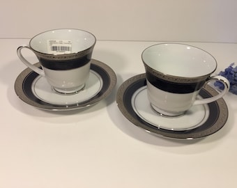 Op de afbeelding: Twee witte porseleinen theekopjes met bijpassende schoteltjes, voorzien van een brede marineblauwe band en een decoratieve zilverkleurige metalen rand. De binnenkant van het linker kopje bevat een kleine witte sticker met zwarte tekst.