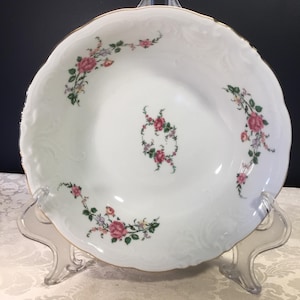 May include: White porcelain serving bowl with a scalloped edge and gold trim. The bowl is decorated with pink and red roses and green leaves. The bowl is displayed on a clear plastic stand.