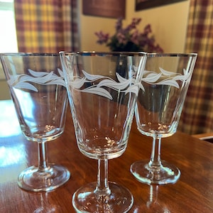May include: Three clear stemmed glasses with a silver rim and a white wave design. The glasses are on a dark wooden surface. The background includes a window, curtains, and a framed picture.