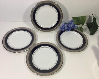 Op de afbeelding: Een set van vier ronde witte porseleinen borden met een brede marineblauwe band en een decoratieve zilverkleurige metalen rand met een reliëfpatroon. De borden zijn in een vierkant opgesteld met een klein boeketje kunstmatige blauwe bloemen.