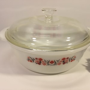 May include: A white casserole dish with a clear glass lid and a clear glass handle. The dish has a floral design in red and pink. The dish is on a white surface.