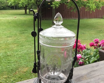 May include: A clear glass jar with an ornate metal lid and stand. The jar has a decorative design and is on a wooden surface.