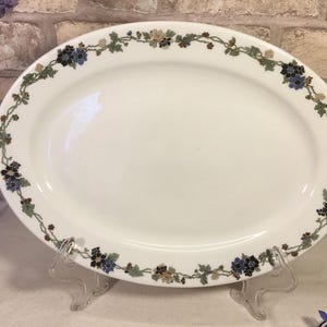 May include: White oval serving platter with a floral border. The border features a repeating pattern of blue, green, and tan flowers and leaves. The platter is displayed on a clear plastic stand.