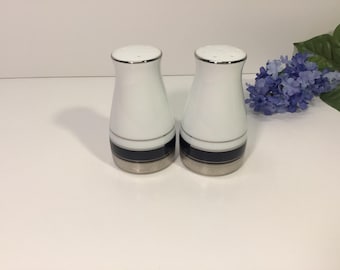Op de afbeelding: Een paar witte zout- en peperstrooiers met een gebogen silhouet, voorzien van een horizontale donkerblauwe band bij de basis en een metallic zilveren rand aan de boven- en onderkant. De strooiers hebben een gladde afwerking en een ontwerp.