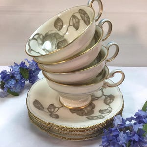 May include: A stack of cream-colored teacups and saucers with a floral design and gold trim. The teacups have a rose motif and are stacked on top of each other. The saucers are stacked below the teacups. The handles are gold-colored.