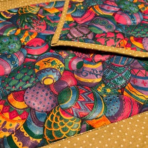 Puede incluir: Un juego de dos manteles individuales acolchados con un patr&oacute;n colorido de huevos de Pascua en tonos de rosa, morado, verde, azul y amarillo. Los manteles individuales tienen un borde dorado con lunares blancos.