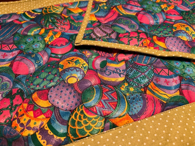 Puede incluir: Un juego de dos manteles individuales acolchados con un patr&oacute;n colorido de huevos de Pascua en tonos de rosa, morado, verde, azul y amarillo. Los manteles individuales tienen un borde dorado con lunares blancos.