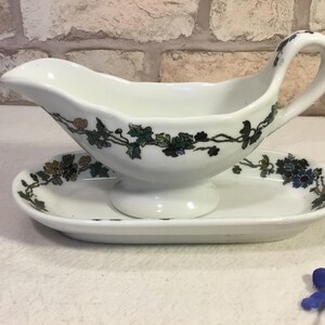 May include: White ceramic gravy boat with a matching oval-shaped saucer. The gravy boat has a handle and spout, and both pieces are decorated with a floral vine pattern in green, blue, and brown.